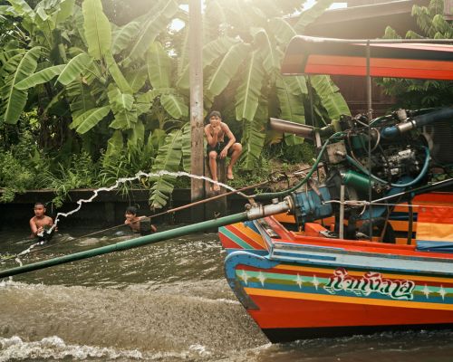 Three young boys swimming in a canal by a colourful boat with a motor, surrounded by lush tropical vegetation on a sunny day.