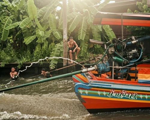 Three young boys swimming in a canal by a colourful boat with a motor, surrounded by lush tropical vegetation on a sunny day.