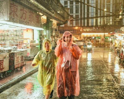 Two smiling women in colorful raincoats walk along a wet city street in heavy rain. Buildings and street vendors are visible in background.