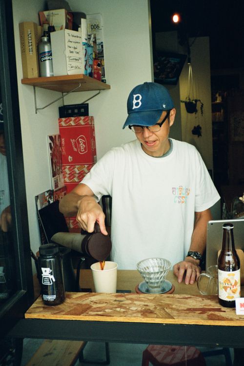 Man wearing a baseball cap and glasses is pouring coffee into a cup at an indoor cafe, with food and drink props displayed on the counter.