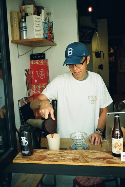 Man wearing a baseball cap and glasses is pouring coffee into a cup at an indoor cafe, with food and drink props displayed on the counter.