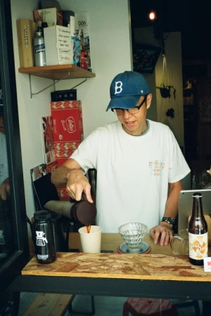Man wearing a baseball cap and glasses is pouring coffee into a cup at an indoor cafe, with food and drink props displayed on the counter.