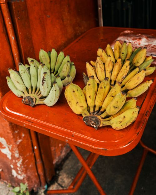 Two bunches of bananas, one yellow and one green, are arranged on a red folding table. The bananas are ripening outdoors.