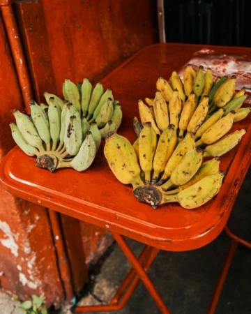 Two bunches of bananas, one yellow and one green, are arranged on a red folding table. The bananas are ripening outdoors.