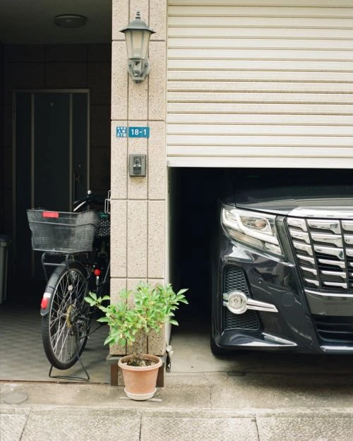 Dark grey car partially parked in a garage with a bicycle and potted plant nearby the entrance of an urban home with neutral tones.