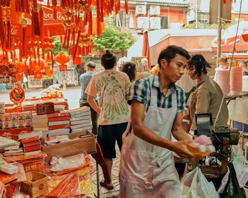 Busy outdoor market scene in Asia with a vendor holding food, surrounded by hanging red lanterns and stacked boxes and packages of goods.