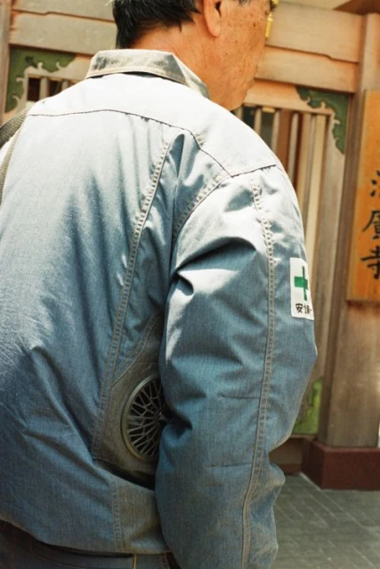 Back view of an adult wearing a blue jacket with a fan and a medical patch near a temple gate with visible Japanese text.