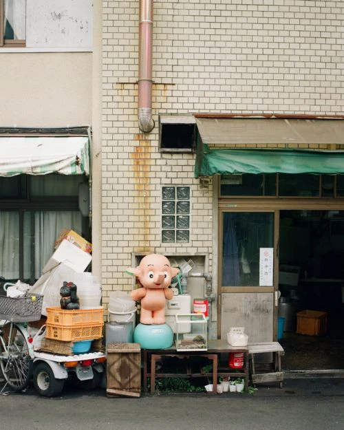 Exterior view features a whimsical pink character figure standing on a blue ball beside a storefront with assorted objects and a bicycle.