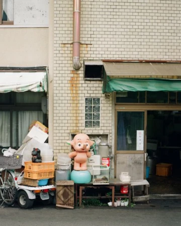 Exterior view features a whimsical pink character figure standing on a blue ball beside a storefront with assorted objects and a bicycle.
