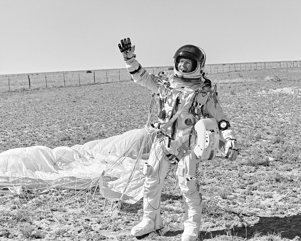 Astronaut in space suit waves hello in an outdoor setting with parachute on the ground and clear sky in a black and white image.