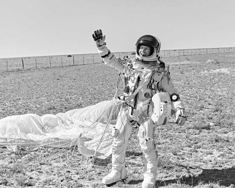 Astronaut in space suit waves hello in an outdoor setting with parachute on the ground and clear sky in a black and white image.