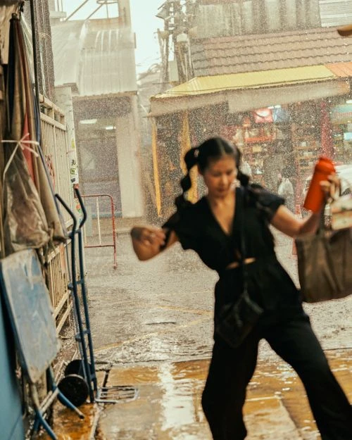 Young woman in black jumpsuit runs through pouring rain in a city street, holding a red water bottle and a tan tote bag. 