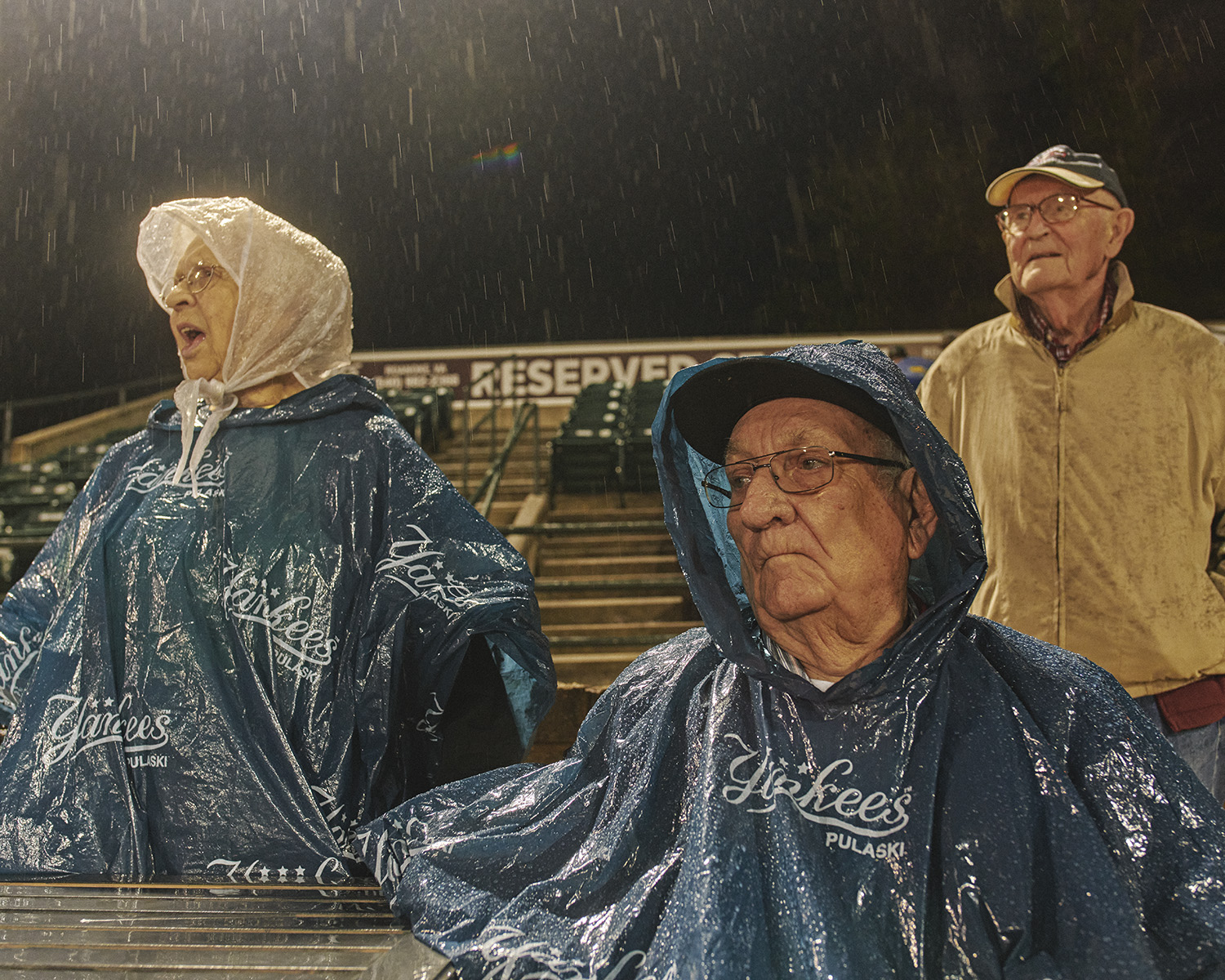 Three elderly baseball fans in ponchos brave the rain at an outdoor game. The woman on the left is animated while the men appear stoic.