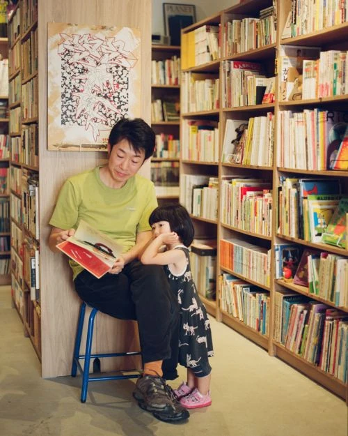 Adult male sits on a blue stool in a book store, reading a book to a young child who is standing next to him.