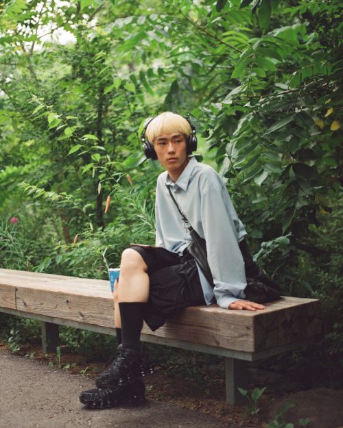 Young adult male with blond hair sits on a wooden bench in a green park, wearing headphones, a blue shirt, and black shorts.