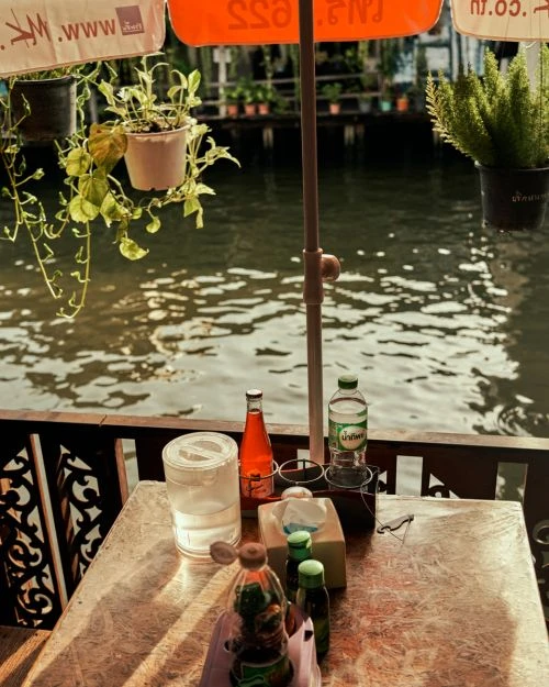 Outdoor dining scene featuring a table with various bottles and condiments next to a waterway, with plants hanging overhead.