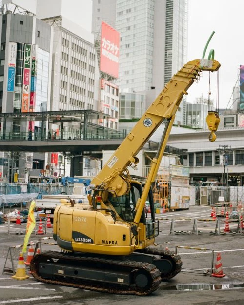 Yellow Maeda mini crane stands on an urban construction site with barriers and tall buildings in the background on an overcast day.