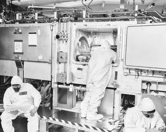 Three technicians in clean suits work inside a bright, sterile industrial facility with pipes and advanced machinery, showcasing technology and precision.