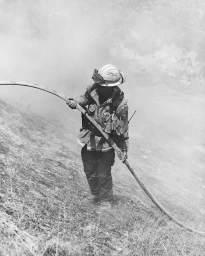 Firefighter in protective gear and helmet holding a hose on a smoky hillside in Los Angeles by Balazs Gardi.
