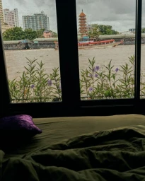 View from inside a room through a window overlooking a wide river with boats, buildings, and a pagoda under an overcast sky.