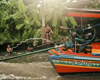 Colorful longtail boat speeding through a canal with children swimming nearby amidst lush tropical vegetation
