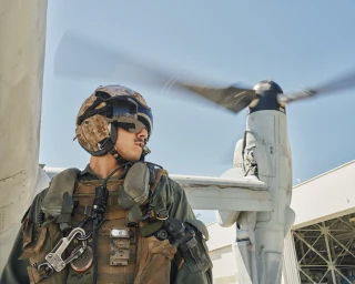 Military aviator in desert camouflage helmet and tactical vest standing before a rotating aircraft propeller