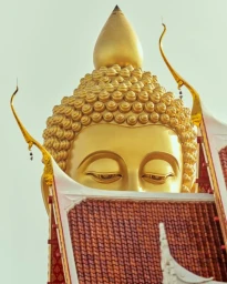 Close-up of a giant golden Buddha statue head positioned directly behind the ornate tiled roof of a temple building.
