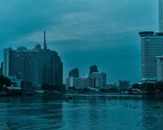 City skyline along a wide river under a deep blue overcast sky by Balazs Gardi