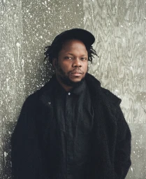 Portrait of a Black man wearing a black cap and dark coat against a weathered textured wall by Balazs Gardi