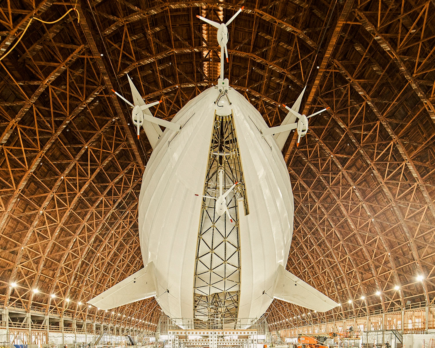 Large white airship with multiple propellers suspended inside a massive wooden hangar with complex truss architecture by Balazs Gardi.