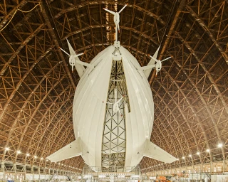 Large white airship with multiple propellers suspended inside a massive wooden hangar with complex truss architecture by Balazs Gardi.
