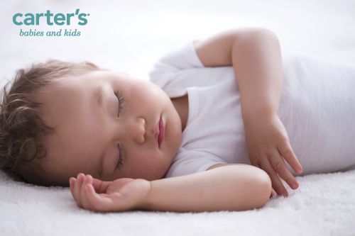 Sleeping baby wearing a white onesie laying on a white textured surface. Studio lit with soft light. Carter's logo is visible.