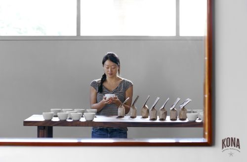 Asian woman at a dark wood table in front of a gray wall, holding a white cup, with tasting cups and coffee bean sample bags lined up.