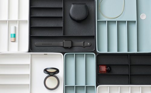 Overhead shot of organized drawer inserts with black, white, and blue compartments containing makeup, watches, and jewelry pieces.