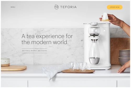 Woman preparing tea with a white tea maker on a bright counter with bowls and glasses in a modern kitchen setting, clean aesthetic.