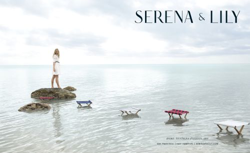 Blonde woman in a white dress stands on a rock formation in a shallow body of water next to several small colorful tables.