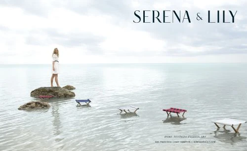 Blonde woman in a white dress stands on a rock formation in a shallow body of water next to several small colorful tables.