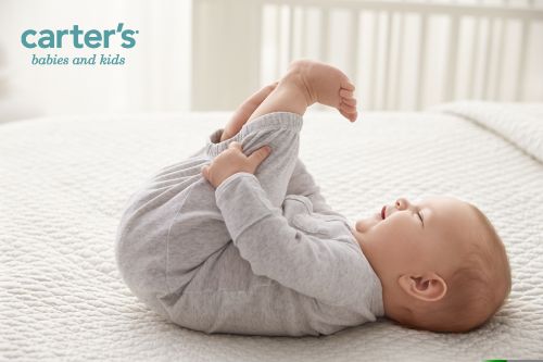 Infant lying on their back on a white quilted blanket, holding their feet and smiling. Carter's logo visible in upper left.