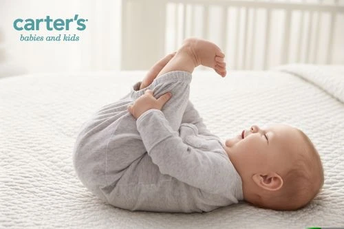 Infant lying on their back on a white quilted blanket, holding their feet and smiling. Carter's logo visible in upper left.