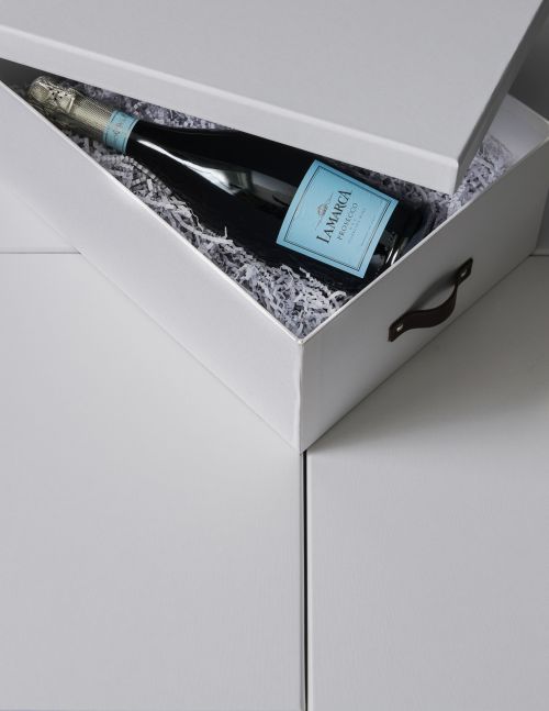 Bottle of La Marca Prosecco inside a white gift box with shredded paper on a white surface. The bottle has a light blue label.