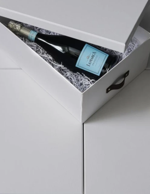 Bottle of La Marca Prosecco inside a white gift box with shredded paper on a white surface. The bottle has a light blue label.