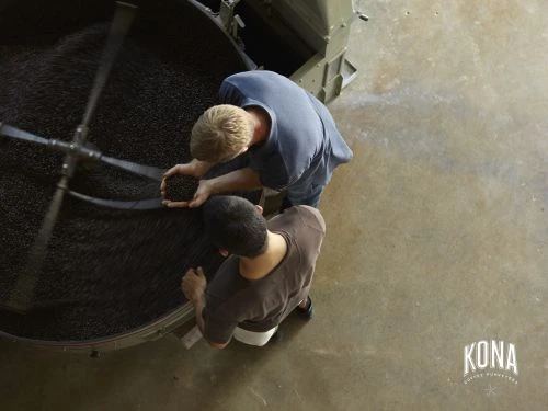 Two workers process coffee beans in a large roaster in an industrial setting, captured from an overhead perspective, featuring the Kona Coffee logo.