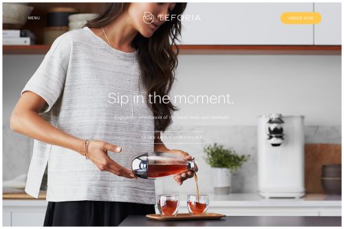Woman pours tea from a glass teapot into two cups on a kitchen counter, with a sleek white tea maker in the background.