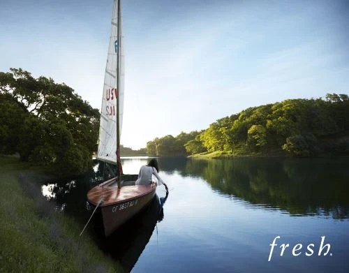 A woman sitting in a vintage sailboat on a lake surrounded by trees, with sunlight creating a serene atmosphere and a sense of leisure.