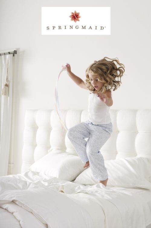 A young girl with curly hair jumps on a bed with white linens. She wears pajamas and is holding a ribbon in a brightly lit room.