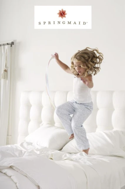 A young girl with curly hair jumps on a bed with white linens. She wears pajamas and is holding a ribbon in a brightly lit room.