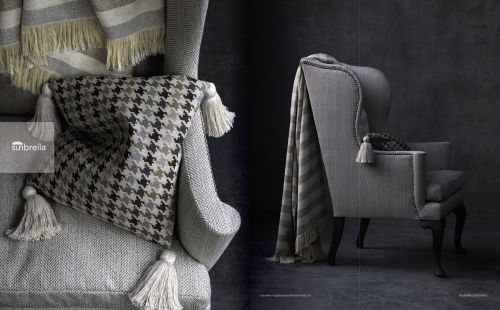 Close-up of a grey armchair with a houndstooth patterned pillow, fringed throw blanket, and tassel details against a dark textured wall