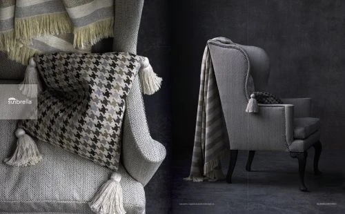 Close-up of a grey armchair with a houndstooth patterned pillow, fringed throw blanket, and tassel details against a dark textured wall