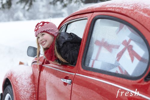 Young girl smiles from the window of a red vintage car filled with white gifts in a snowy winter landscape. Holiday cheer.