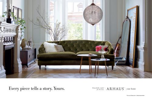 Interior view of a living room featuring an olive green tufted sofa, white walls, wood flooring, and decorative elements like a chandelier and mirror.